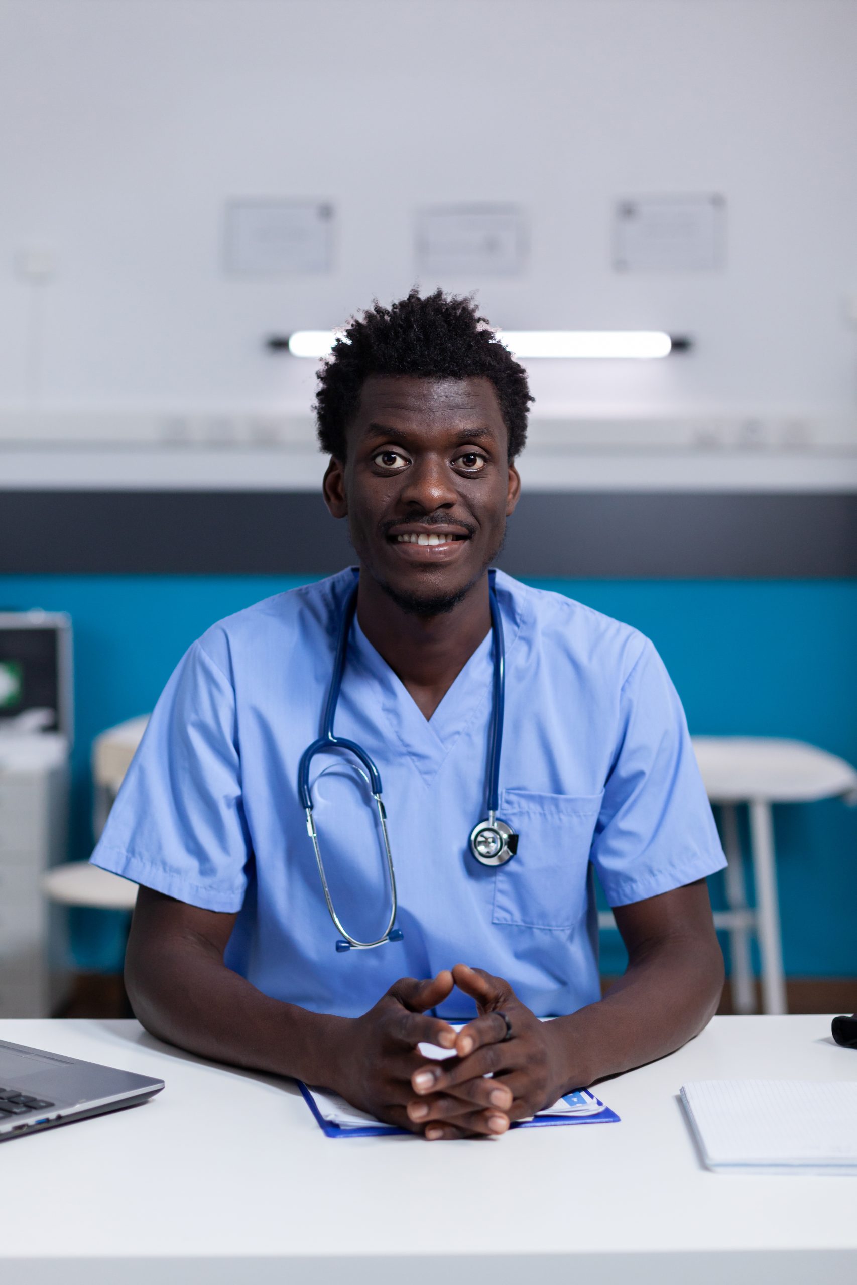 Portrait of african american man with nurse profession smiling looking at camera in healthcare cabinet. Black adult sitting at desk with medical instruments and laptop for consultation