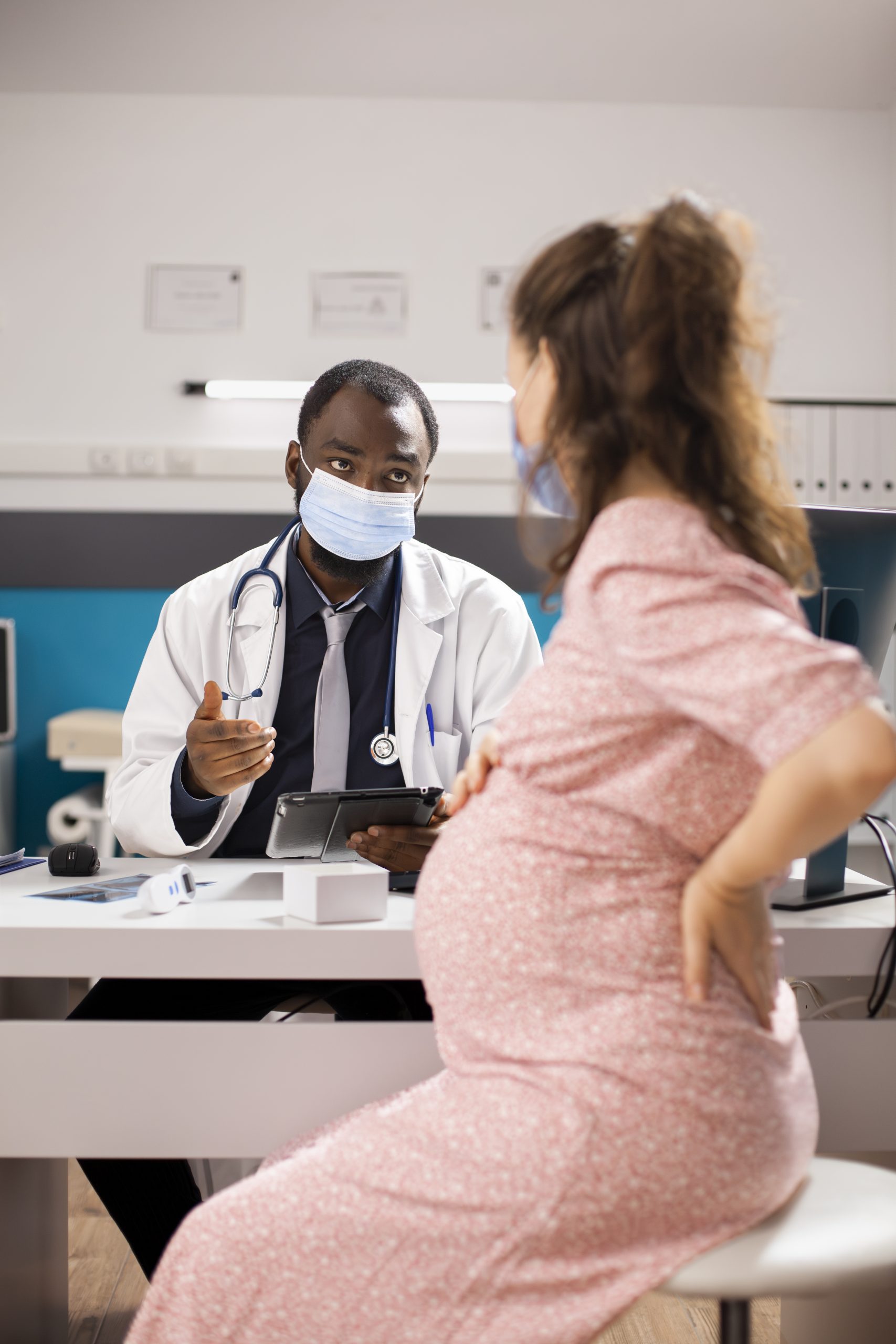Male obstetrician discusses pregnancy care with white woman during checkup visit. Black doctor uses digital tablet to show information to pregnant lady about prescription medicine and health plan.